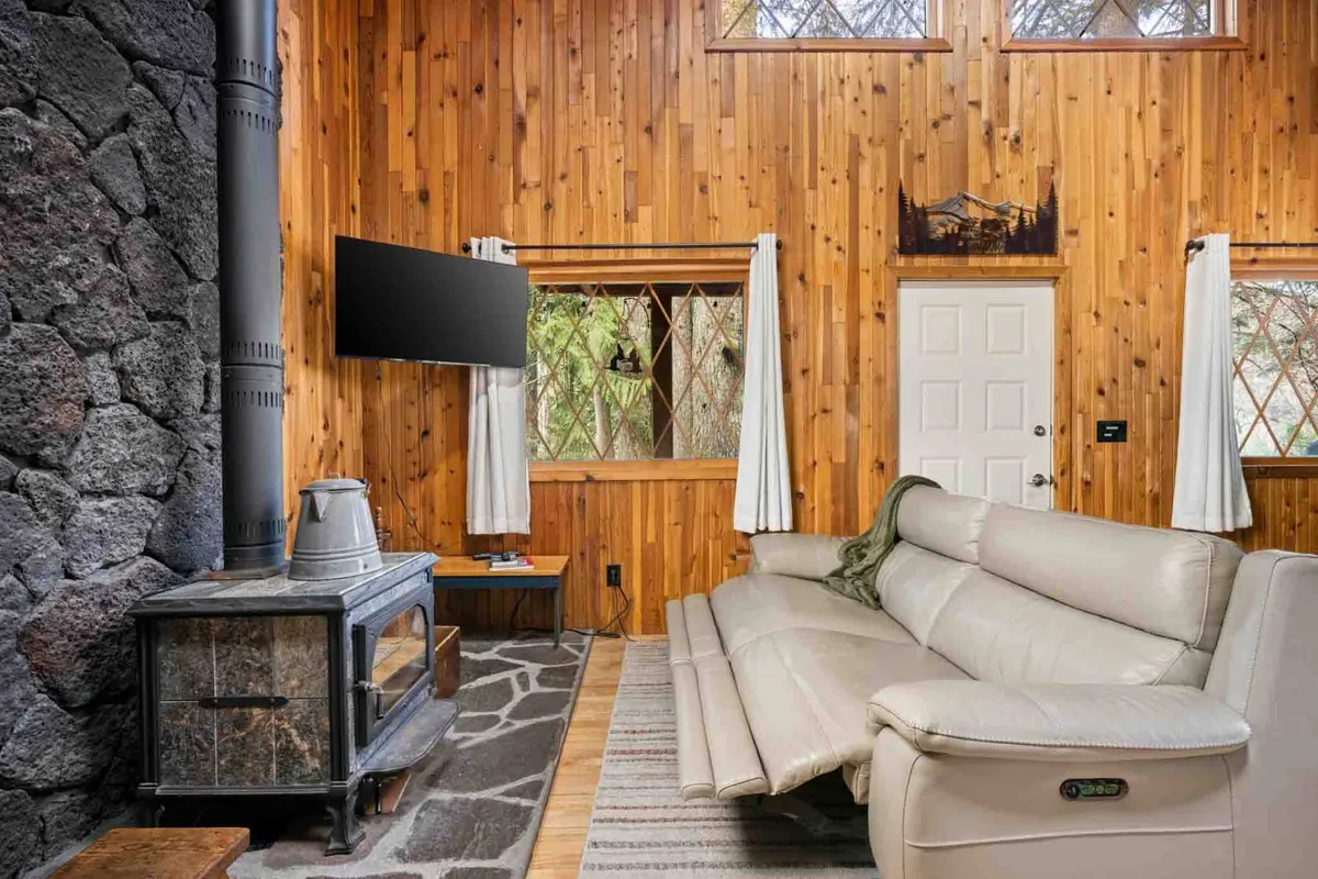 A spacious main gathering area with soaring ceilings, a stone fireplace, a leather sofa, and expansive windows that bring the outdoors in. The room is filled with natural light and framed by rich wooden walls, capturing the essence of mountain cabin living.