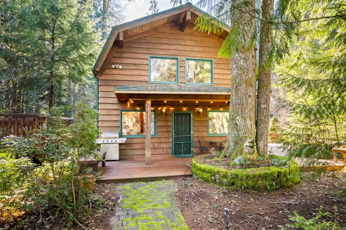 This image showcases the wooden facade of Crystal Creek Cabin, accented by green windows and a welcoming front porch surrounded by lush trees. The scene hints at relaxing on the porch, barbecuing, or simply enjoying the natural setting with family and friends.