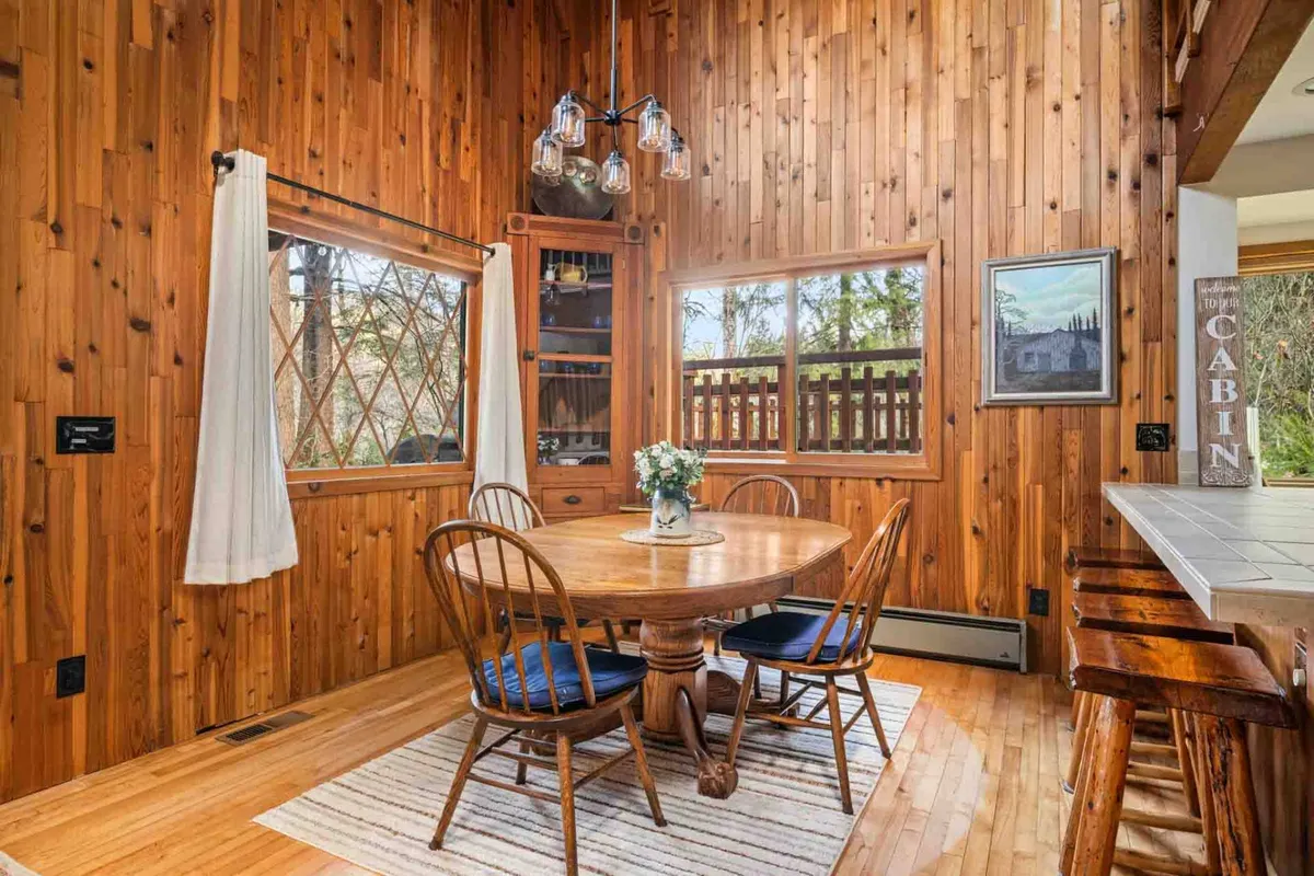 A charming dining area featuring a sturdy wooden table, elegant light fixtures, large windows allowing daylight to fill the space, and decorative shelving. The setup invites intimate family meals and gatherings with friends.