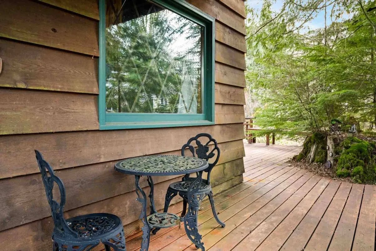 An inviting wooden deck with a small round table and ornate chairs, set amidst lush greenery. The deck overlooks a peaceful, natural setting, blending indoor comfort with the outdoors—ideal for relaxing morning coffee or quiet moments with furry friends after a walk.