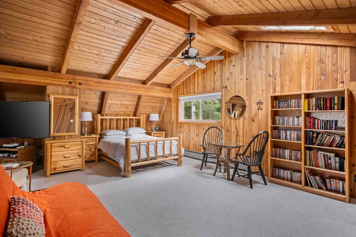 A tranquil bedroom with a wooden bed, large window letting in natural light, a bookshelf filled with books, and a cozy sitting area with chairs and a table. The space feels peaceful and designed for unwinding, with touches that invite relaxation and reading.