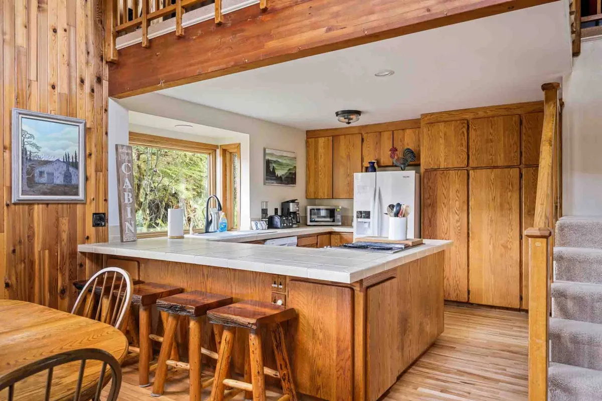 This image features a spacious kitchen with warm wooden cabinetry, a central island, large sunlit window, and stools. The adjacent dining table is in clear view, suggesting a seamless flow between cooking and dining spaces. The room is filled with natural light, enhancing its homey and inviting atmosphere.