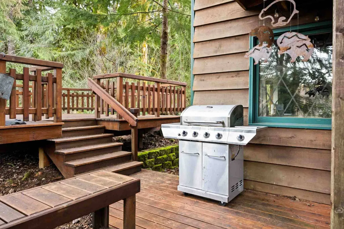 This image showcases a spacious wooden deck surrounded by lush greenery, featuring a grill and ample open space. The setting is perfect for barbecuing, entertaining guests, and enjoying outdoor dining against a relaxed, natural backdrop.
