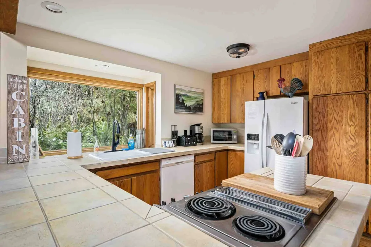 A well-lit kitchen setting with a central island stovetop, ample wooden cabinetry, large window drawing in natural light, and decorative shelves. This space feels both functional and welcoming, ideal for meal preparation and group cooking.
