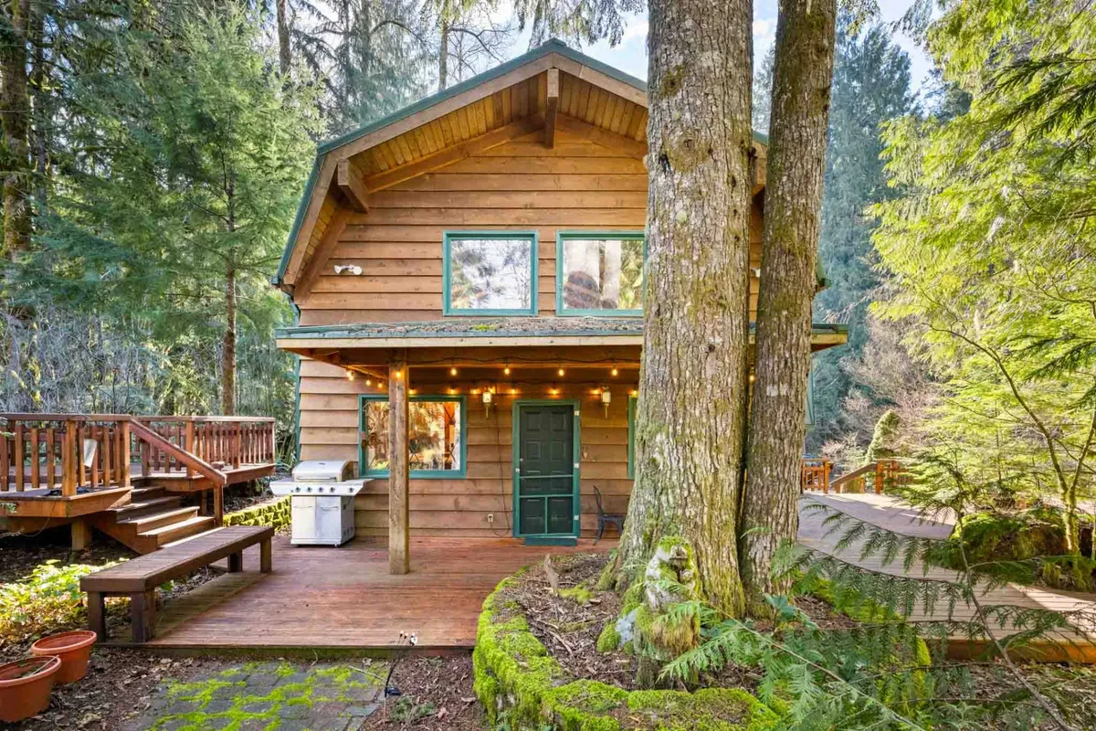 The image features a wooden cabin nestled among large trees, with a prominent deck area and a grill. The scene is warm and welcoming, inviting guests to relax on the deck, stargaze, or gather for early morning coffee surrounded by nature.