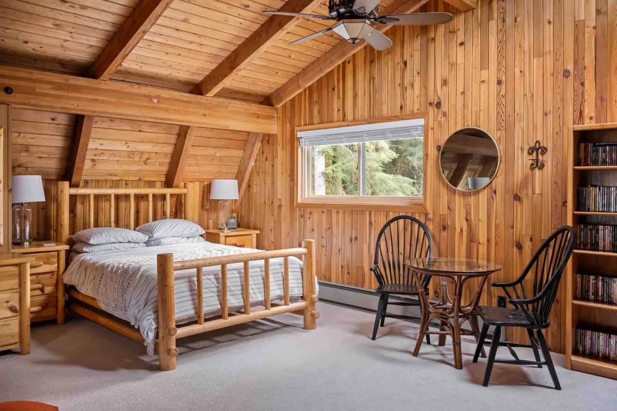 A cozy bedroom featuring a wooden bed frame and accent wall, with a round mirror and a window offering a view of the outdoors. The room is warmly lit and decorated in a mountain-inspired style, creating an inviting and restful atmosphere.