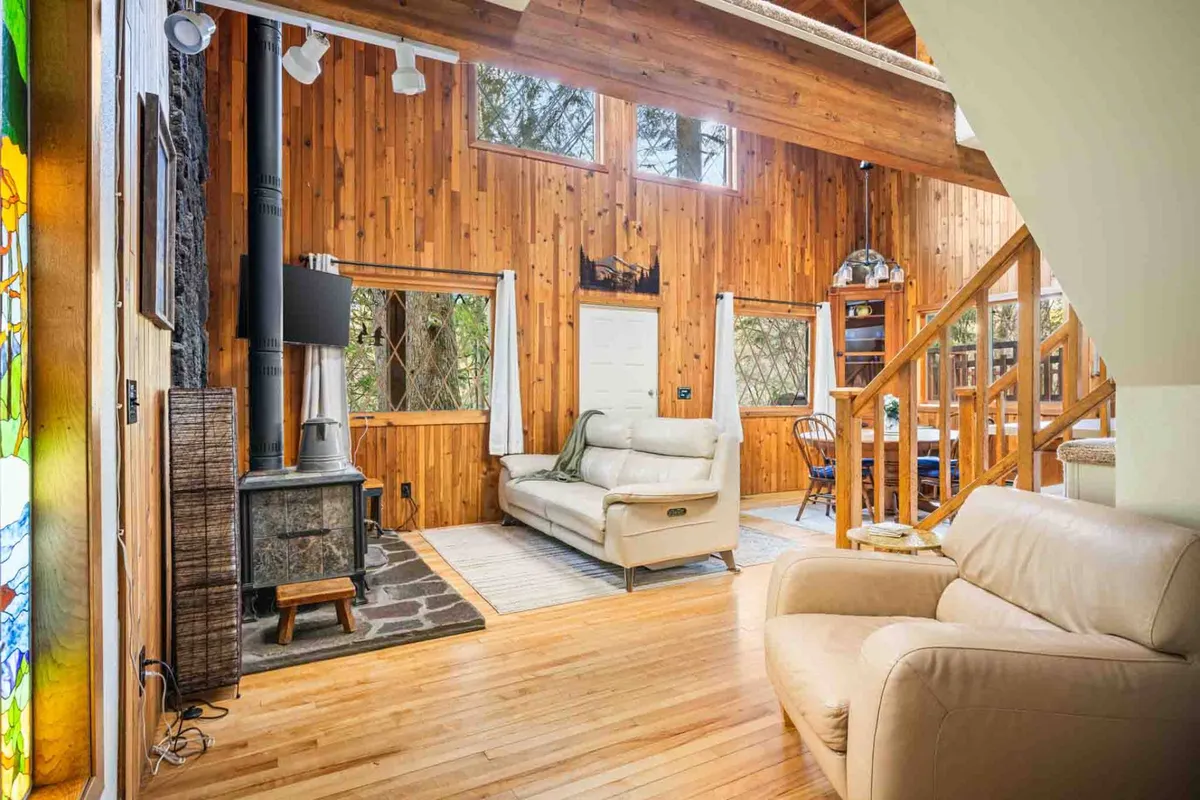 A cozy interior with rich wooden walls, plush seating arranged around a wood-burning stove, and a dining area bathed in natural light from large windows that reveal glimpses of the surrounding forest. The space feels both intimate and inviting, perfect for family gatherings or quiet moments by the fire.