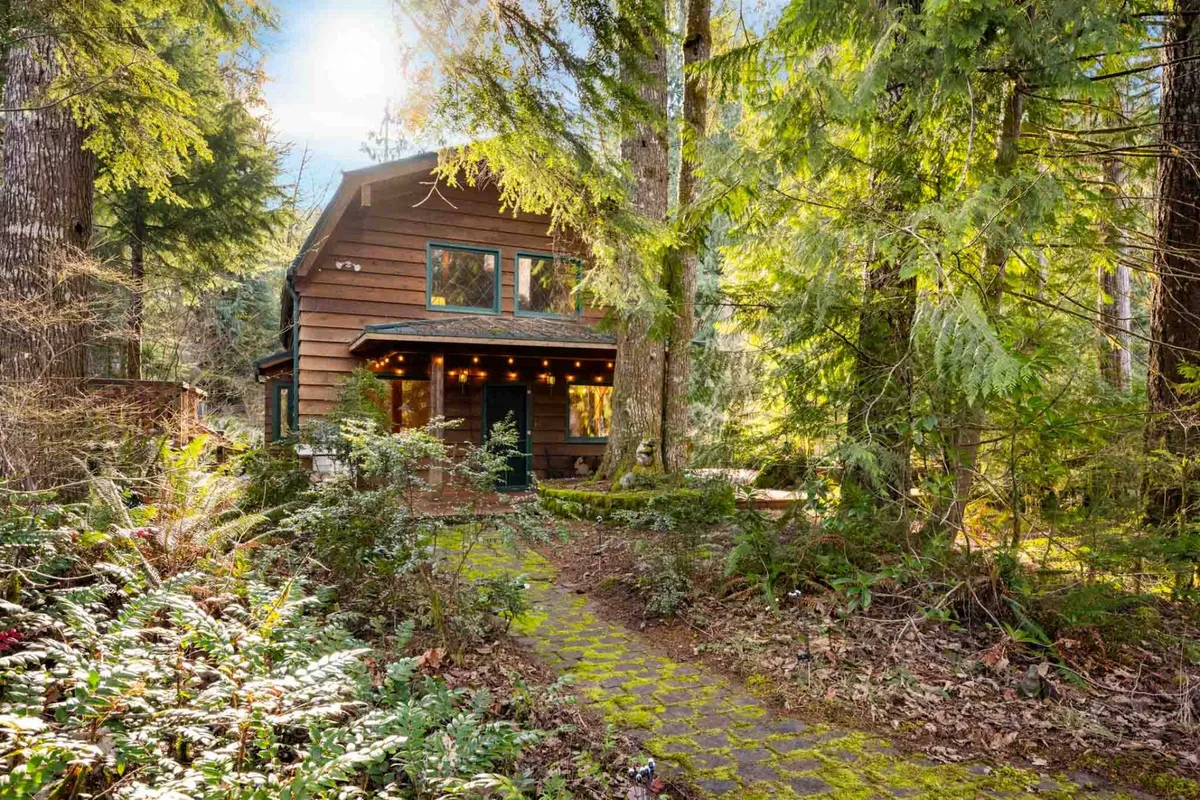 A welcoming scene of the wooden cabin nestled among dense greenery, with a visible pathway, warm window lighting, and decorative outdoor lights. The inviting exterior suggests easy access to nature trails and relaxing evenings outdoors.