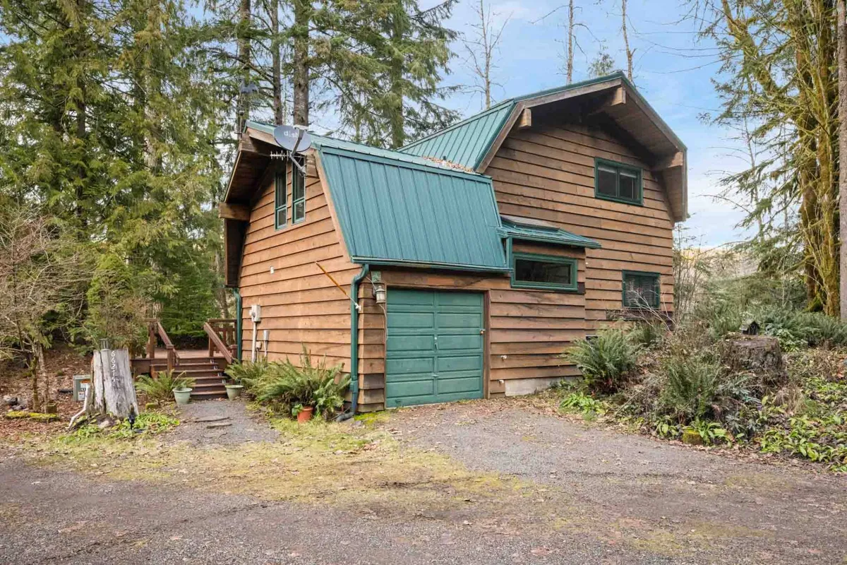 This image showcases the wooden exterior of the property nestled among tall trees, with a green metal roof and a driveway. The outdoor setting is inviting and showcases ample space for outdoor activities, such as nature walks and gatherings with pets. The cozy mood is accentuated by golden sunset lighting and the tranquil natural surroundings.