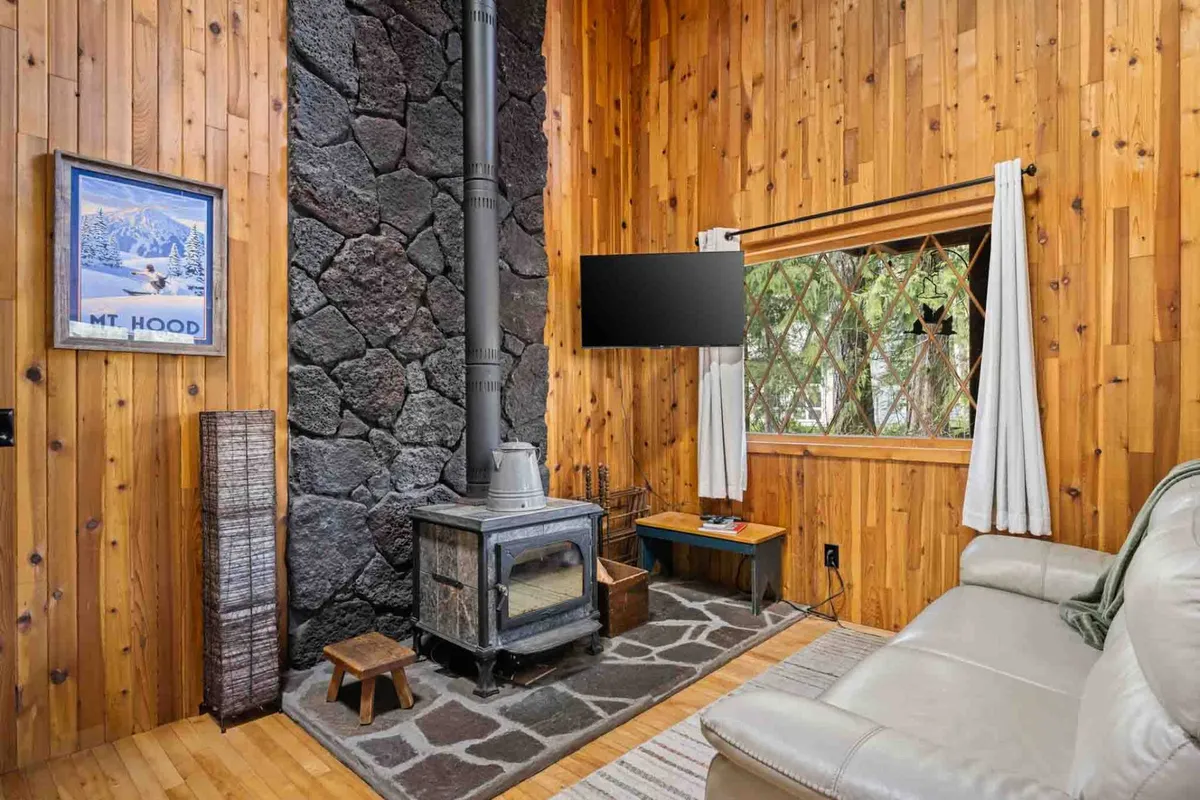 A cozy living room featuring a stone fireplace, rustic wooden walls, a plush leather couch, and large windows that let in ample natural light. Framed mountain artwork and forest views through the windows further enhance the inviting, serene environment.